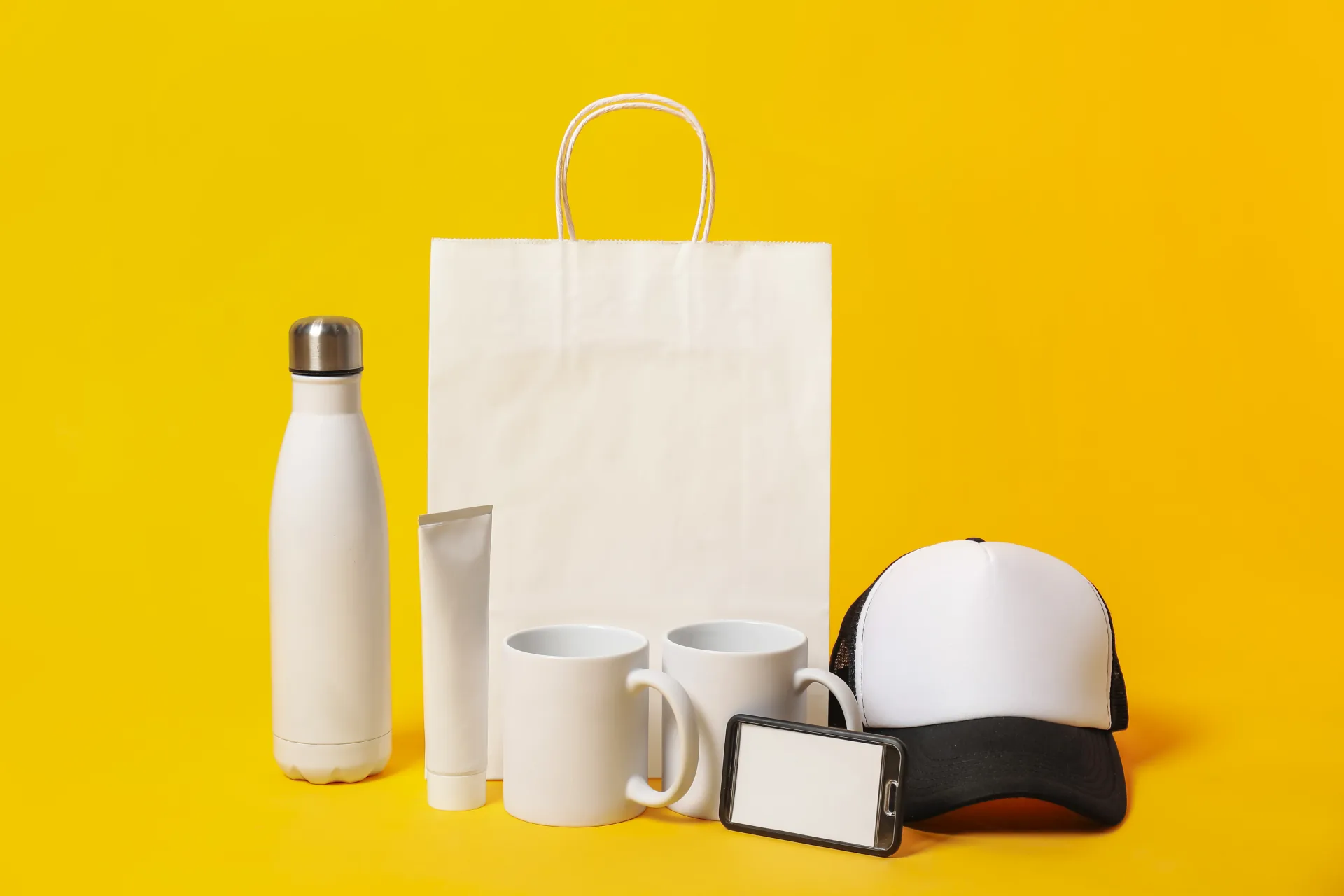 Blank mugs, bottle, cap, bag and phone arranged for custom branding on a yellow background